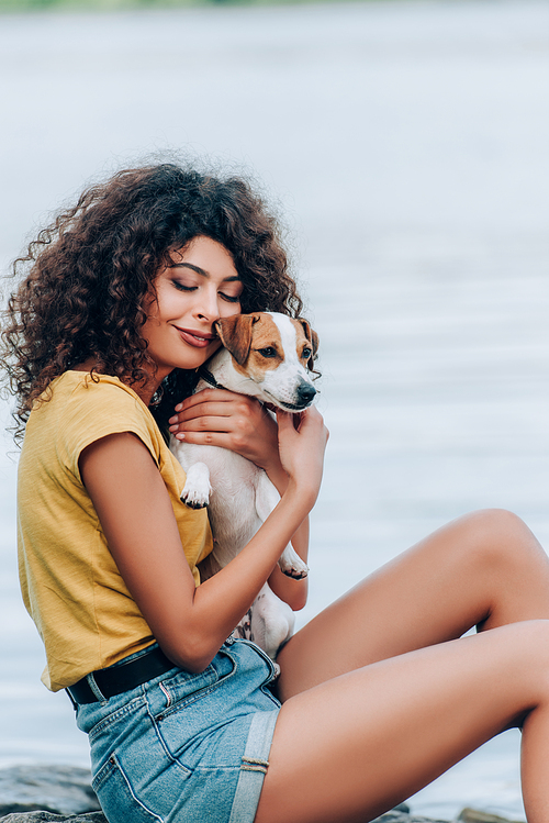 young woman in summer outfit cuddling jack russell terrier dog with closed eyes near lake