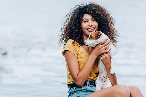 excited, curly woman in summer outfit embracing jack russell terrier dog near lake