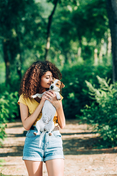 young, curly woman in summer outfit kissing jack russell terrier dog in park