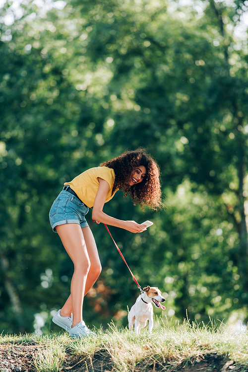 young woman in summer outfit laughing while taking photo of jack russell terrier dog in park