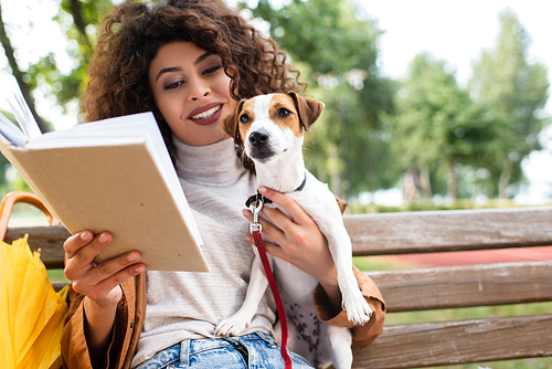 joyful woman reading book and holding jack russell terrier dog on bench in park