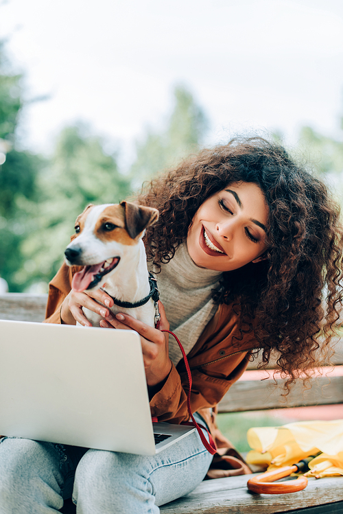 joyful freelancer cuddling jack russell terrier dog while sitting on bench with laptop