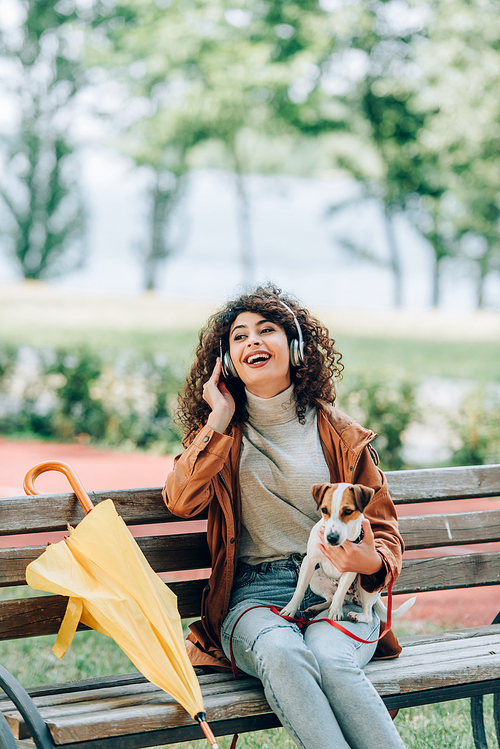 laughing woman in wireless headphones looking away while sitting in park with jack russell terrier dog near yellow umbrella