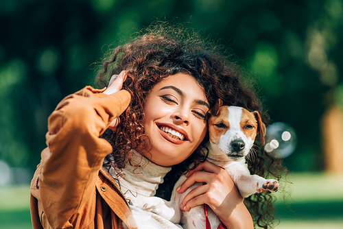 Selective focus of young curly woman with hand near head holding jack russell terrier outdoors