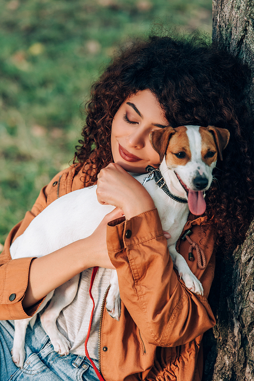 Young woman in raincoat hugging jack russell terrier near tree