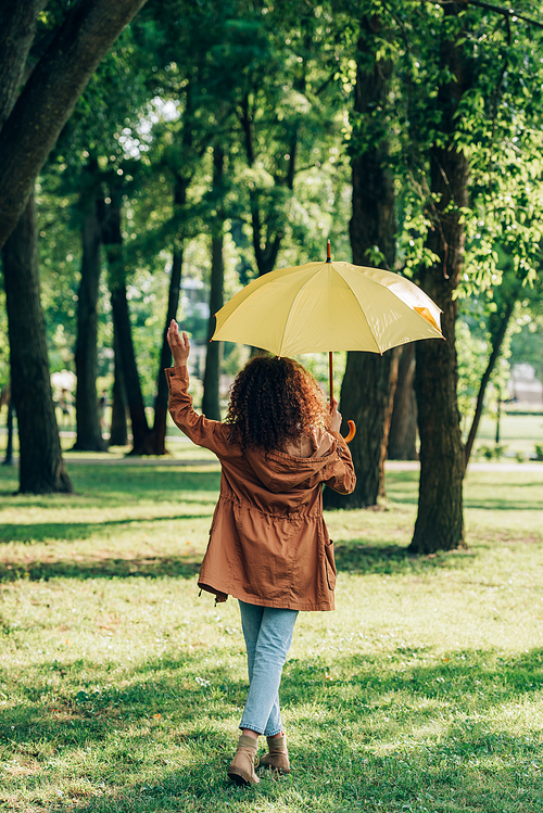 Back view of young woman in autumn outfit holding umbrella while walking in park