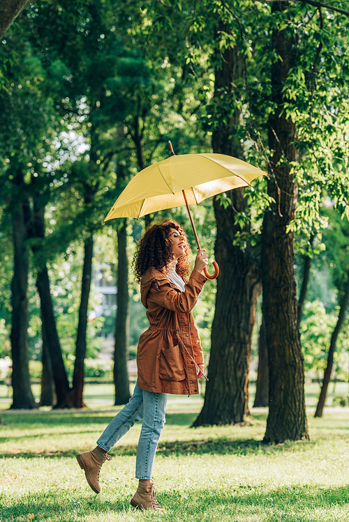 Side view of curly woman in raincoat and jeans holding umbrella in park