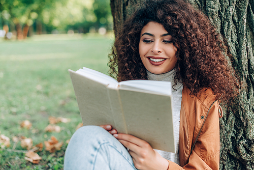 Selective focus of curly woman in autumn outfit reading book in park