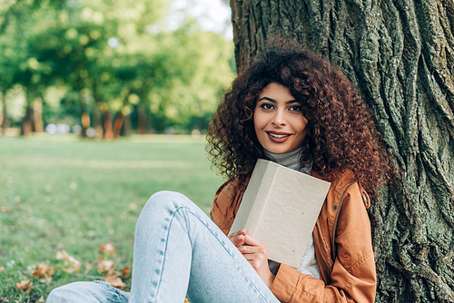 Selective focus of curly woman in raincoat holding book near tree in park