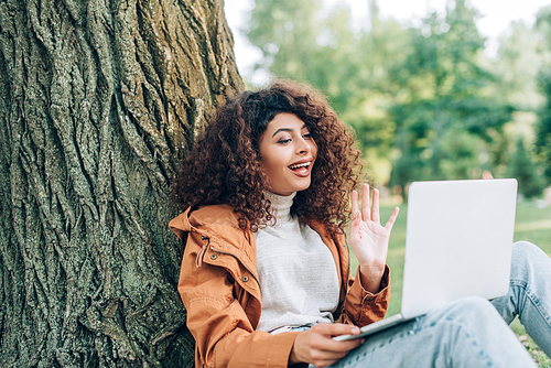 Selective focus of woman in raincoat having video call on laptop in park