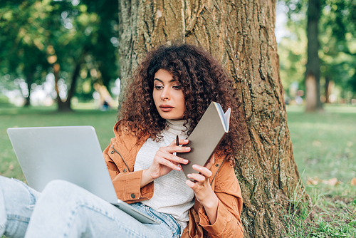 Selective focus of woman looking at laptop while holding notebook and pen near tree in park