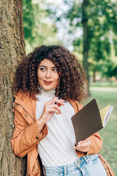 Selective focus of curly woman in raincoat looking away while holding pen and notebook in park