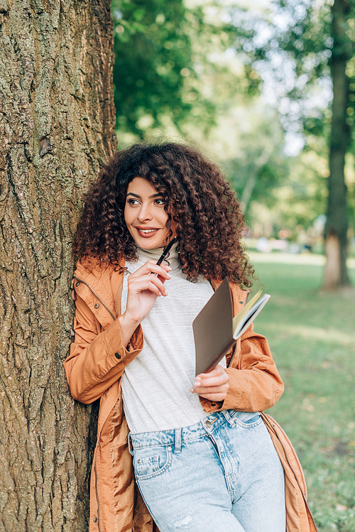 Curly woman in raincoat holding pen and notebook near tree in park