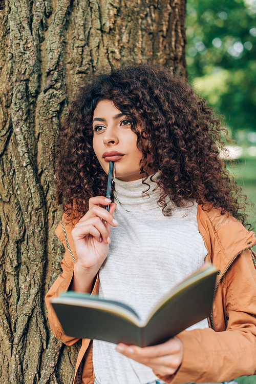 Selective focus of pensive woman in autumn outfit holding pen and notebook in park
