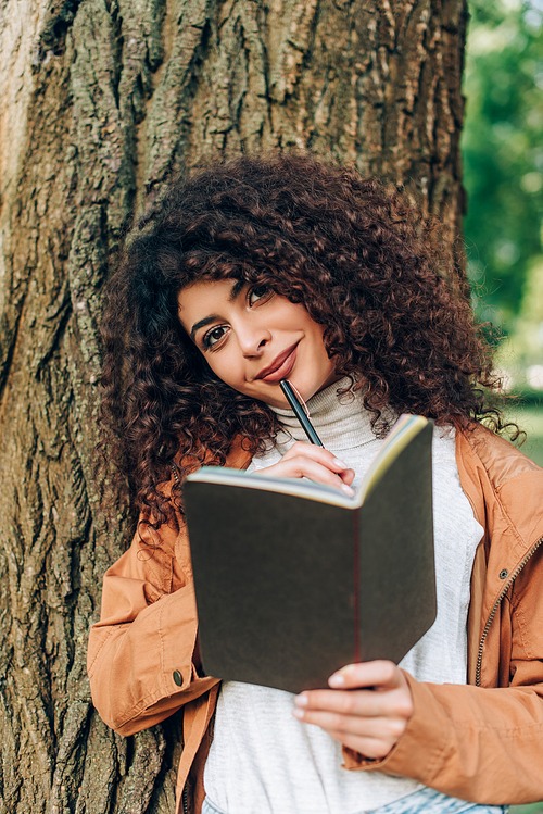 Selective focus of young woman in raincoat holding pen and notebook while  near tree