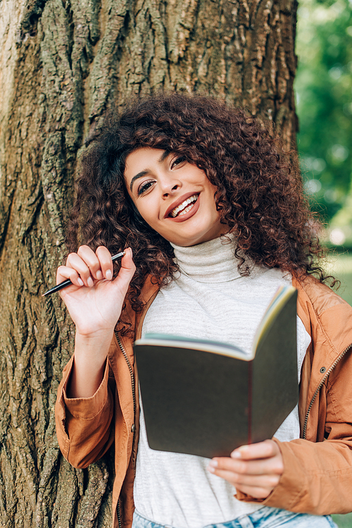 Selective focus of curly woman with notebook and pen  near tree
