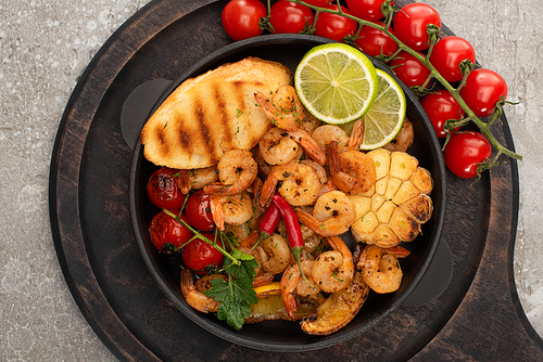 top view of fried shrimps with grilled toasts, vegetables, cherry tomatoes and lime on wooden board on grey concrete background