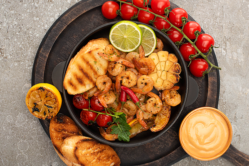 top view of fried shrimps with grilled toasts, vegetables, cherry tomatoes and lime on wooden board with sauce on grey concrete background