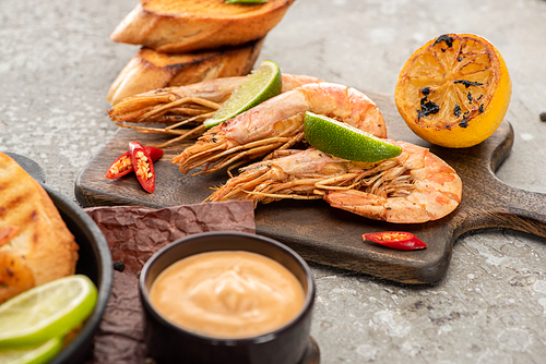 selective focus of fried shrimps with grilled toasts, lemon and lime on wooden board near sauce on grey concrete background