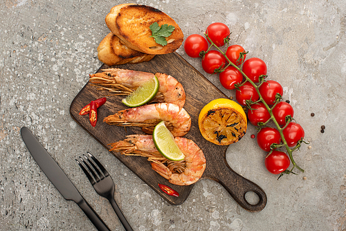 top view of fried shrimps with grilled toasts, lemon and lime near cherry tomatoes on wooden board near cutlery on grey concrete background