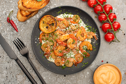 top view of fried shrimps in sauce with dill and lemon near cutlery, cherry tomatoes and toasts on grey concrete background