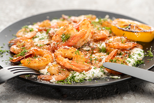 selective focus of fried shrimps in sauce with dill and lemon in black plate on grey concrete background with cutlery