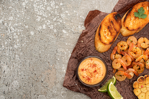top view of fried shrimps with sauce, grilled toasts, lime on parchment paper on grey concrete background