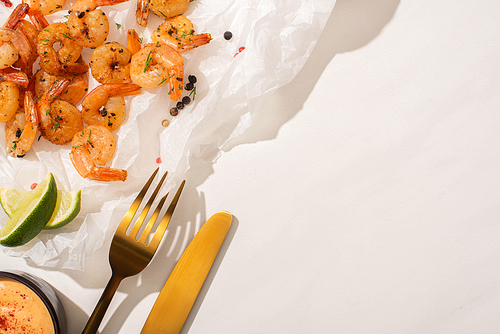 top view of fried shrimps on parchment paper with cutlery, pepper, sauce and lime on white background