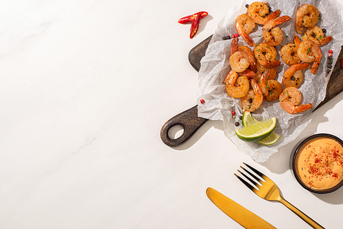 top view of fried shrimps on parchment paper on wooden board with cutlery, chili pepper, sauce and lime on white background