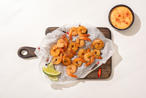 top view of fried shrimps on parchment paper on wooden board with chili pepper, sauce and lime on white background