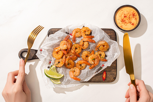 cropped view of woman eating fried shrimps on parchment paper on wooden board with chili pepper, sauce and lime on white background
