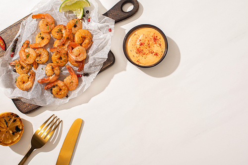 top view of fried shrimps on parchment paper on wooden board near grilled lemon, sauce and cutlery on white background