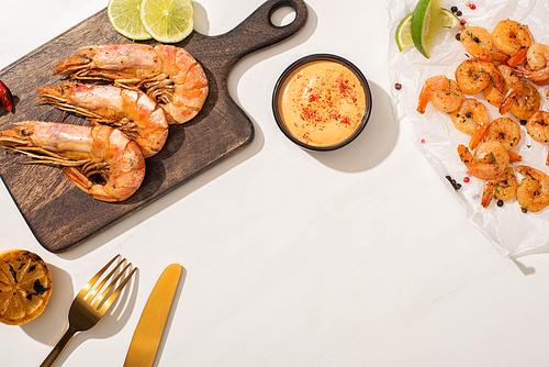top view of fried shrimps on parchment paper and wooden board near grilled lemon, sauce and cutlery on white background