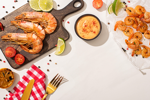 top view of fried shrimps on parchment paper and wooden board with cutlery, plaid napkin, vegetables, sauce and lime on white background