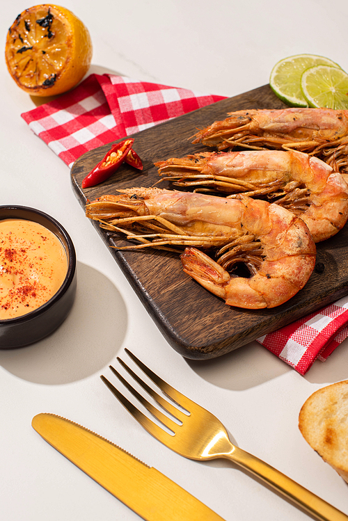 fried shrimps on wooden board on plaid napkin with sauce and lime near golden cutlery on white background
