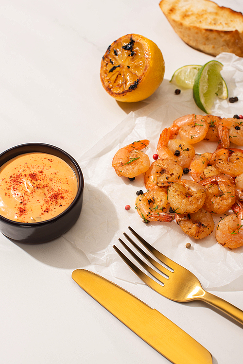 fried shrimps on parchment paper with sauce and lime near golden cutlery on white background