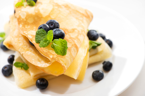 close up view of tasty crepes with mint and blueberries on plate on white background