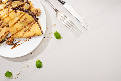 top view of tasty crepes with chocolate spread and walnuts on plate near cutlery and mint leaves on grey background