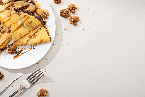 top view of tasty crepes with chocolate spread and walnuts on plate near cutlery on grey background