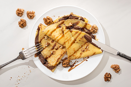 top view of tasty crepes with chocolate spread and walnuts on plate with cutlery on grey background