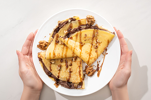 cropped view of woman holding tasty crepes with chocolate spread and walnuts on plate on grey background
