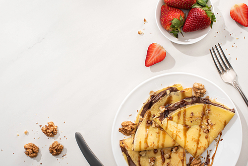 top view of tasty crepes with chocolate spread and walnuts on plate near bowl with strawberries and cutlery on grey background