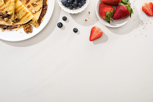 top view of tasty crepes with chocolate spread and walnuts on plate near bowls with blueberries and strawberries on grey background