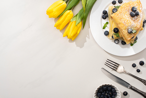 top view of tasty crepes with blueberries and mint on plate near yellow tulips and cutlery on grey background