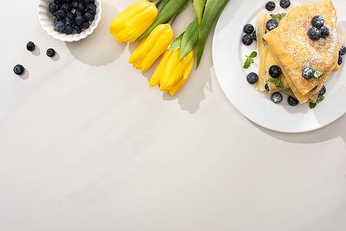 top view of tasty crepes near bowl of blueberries and yellow tulips on grey background