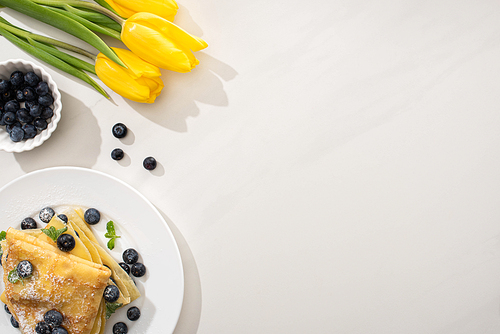 top view of tasty crepes with blueberries and mint on plate near yellow tulips on grey background