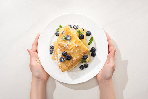 cropped view of woman holding tasty crepes with blueberries and mint on plate on grey background