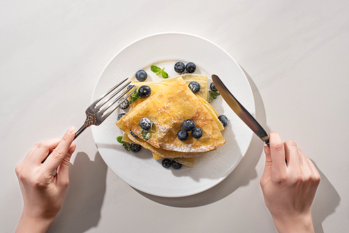 cropped view of woman eating tasty crepes with blueberries and mint on grey background