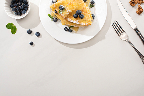 top view of tasty crepes with blueberries and mint on plate near walnuts and cutlery on grey background