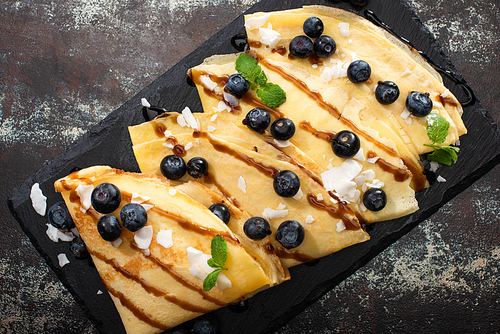 top view of tasty crepes with blueberries, mint and coconut flakes served on board on textured background